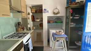 Une cuisine compacte équipée d’une cuisinière à gaz, d’un four et d’un lave-linge. On y trouve une petite table recouverte d’une nappe bleue et un tabouret. La cuisine est dotée de meubles en bois et d’un sol carrelé.