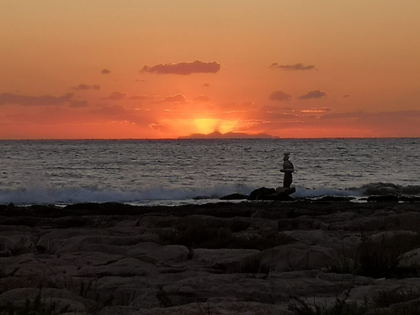 Un coucher de soleil sur une côte rocheuse, avec une personne debout sur les rochers.