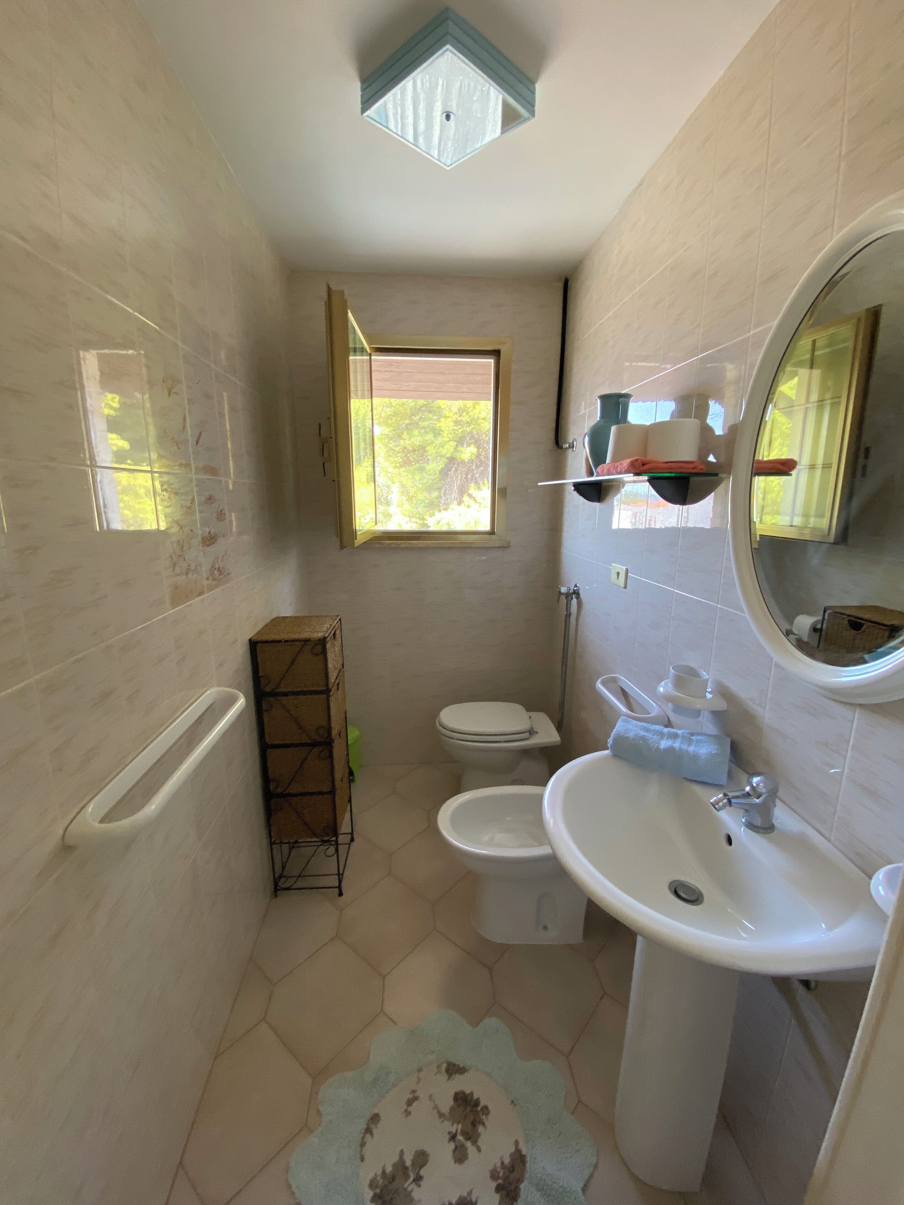 Een kleine badkamer met toilet, bidet en wastafel. Er is een raam dat voor daglicht zorgt en een spiegel boven de wastafel.