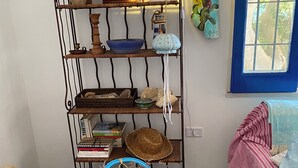 Une pièce avec une étagère en métal où sont disposés divers objets décoratifs, une chaise en osier avec un panier tressé, et une fenêtre donnant sur un paysage verdoyant.