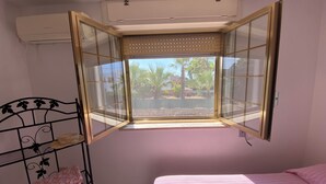 Une chambre à coucher avec un lit, une fenêtre donnant sur des palmiers, une étagère en métal et un climatiseur.