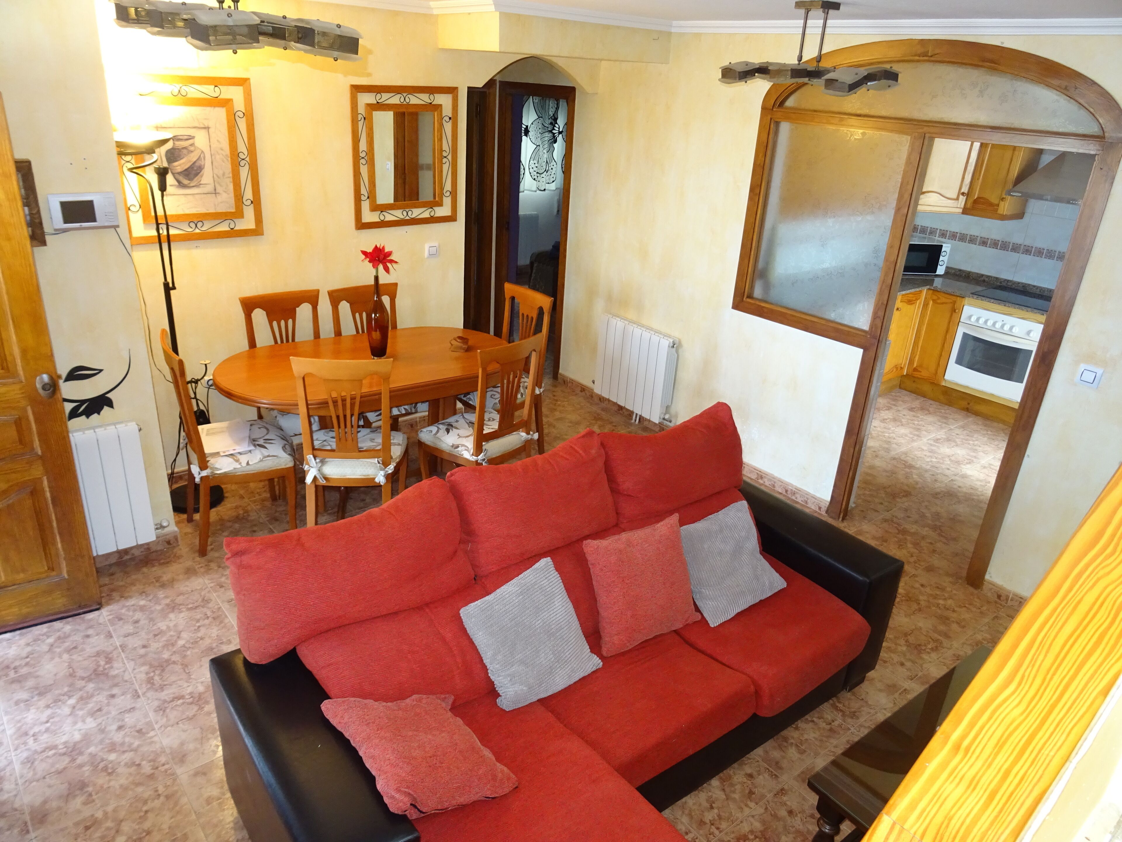 A living room with a red sofa, dining table, chairs, and a kitchen area visible through an archway.