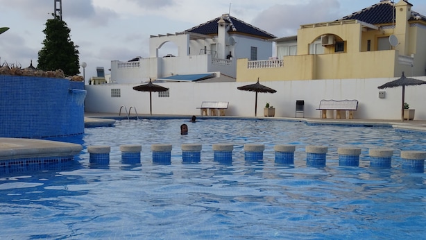 Piscina con agua azul cristalina, rodeada por un muro blanco con azulejos azules. Se ven varios sombrillas y una silla de salvavidas.