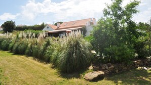 Une maison avec un toit recouvert de tuiles rouges, entourée de hautes herbes et d’un mur de pierre.
