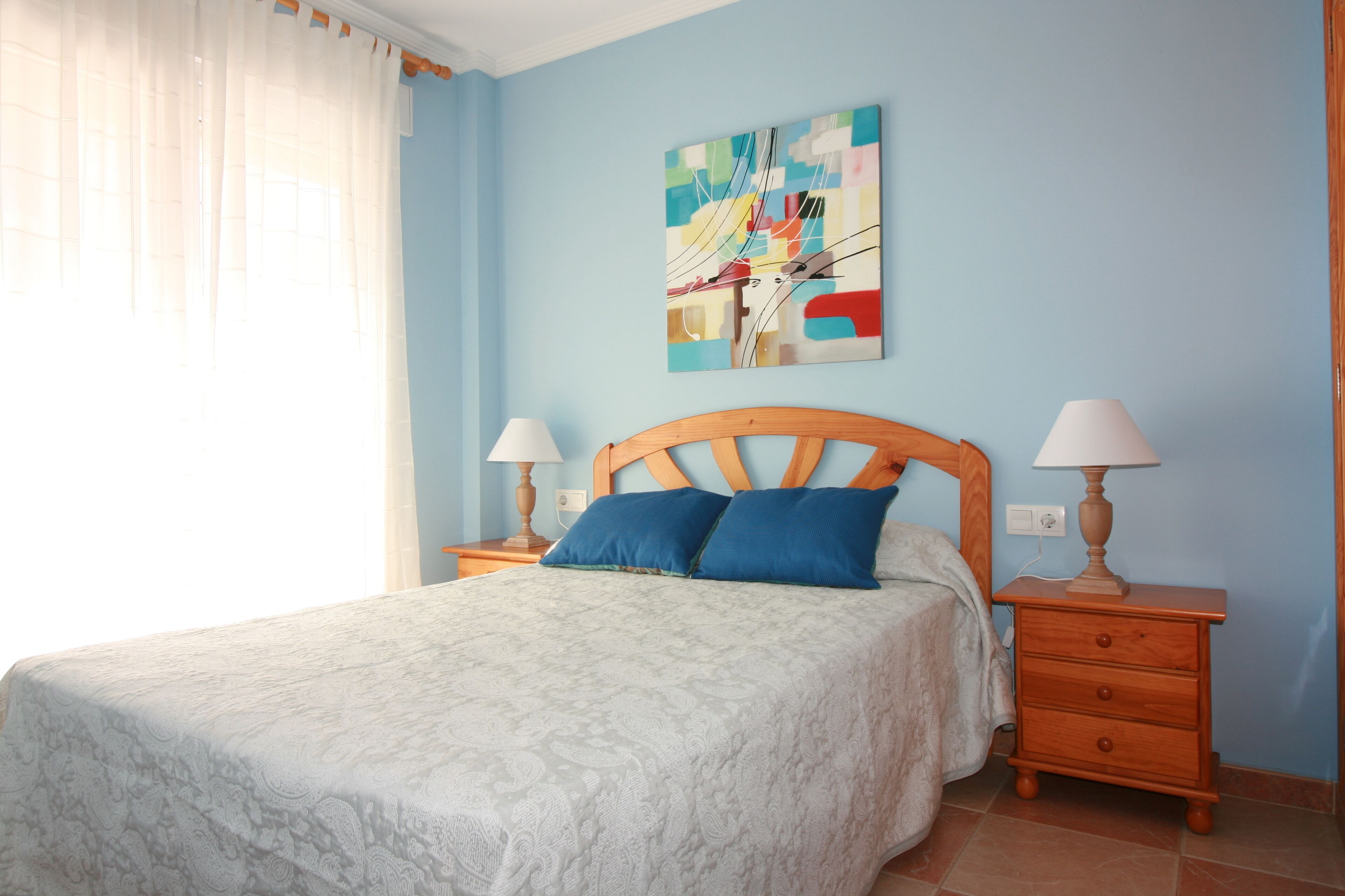 Un dormitorio con una cama, dos mesitas de noche, una mesilla auxiliar, un cuadro colgado en la pared y una ventana con cortinas.