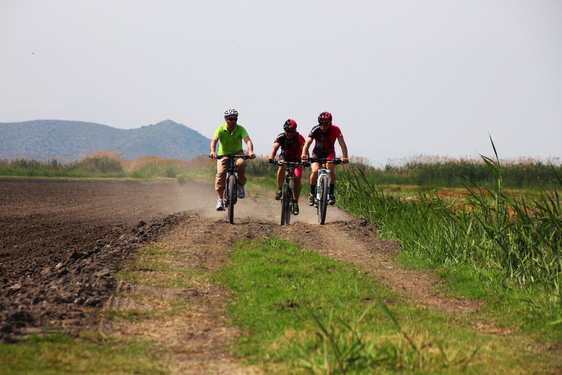 Trois cyclistes sur un chemin de terre, avec de l’herbe de chaque côté et une montagne en arrière-plan.