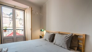 Quarto com cama, mesa de cabeceira, abajur e vista para prédios pela janela.