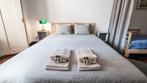 Cama de casal com colcha cinza, duas almofadas e duas toalhas dobradas, com artigos de higiene pessoal em uma cabeceira de madeira.