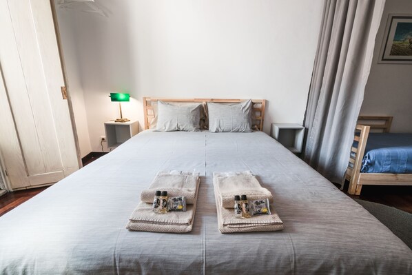 Un lit double avec une couvre-lit grise, deux oreillers et deux serviettes pliées, le tout posé sur une tête de lit en bois, avec des articles de toilette à proximité.