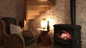 Une pièce chaleureuse avec un mur en pierre, un escalier en bois, un fauteuil en osier, une table avec une lampe et un poêle à bois.
