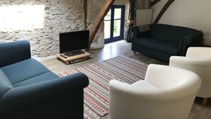 Un salon chaleureux avec un mur en pierre, des poutres apparentes, un téléviseur posé sur un meuble en bois et un tapis à motifs.