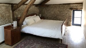 Une chambre avec un plafond en bois, un mur en pierre, un lit recouvert de draps blancs, une table de chevet, une commode en bois et un tapis à motifs.
