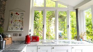 A kitchen with a large window, a red kettle, and a flowered wall hanging. - Lovely property in the heart of Somerset (Wells)