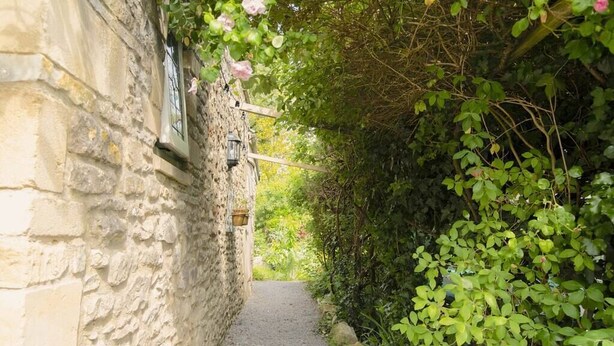 A narrow stone pathway flanked by lush greenery and a stone wall with a window and wall-mounted light fixture. - Lovely property in the heart of Somerset (Wells)
