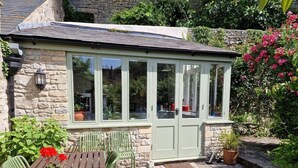 A conservatory with stone walls and a green door, surrounded by potted plants and a wooden table with chairs. - Lovely property in the heart of Somerset (Wells)