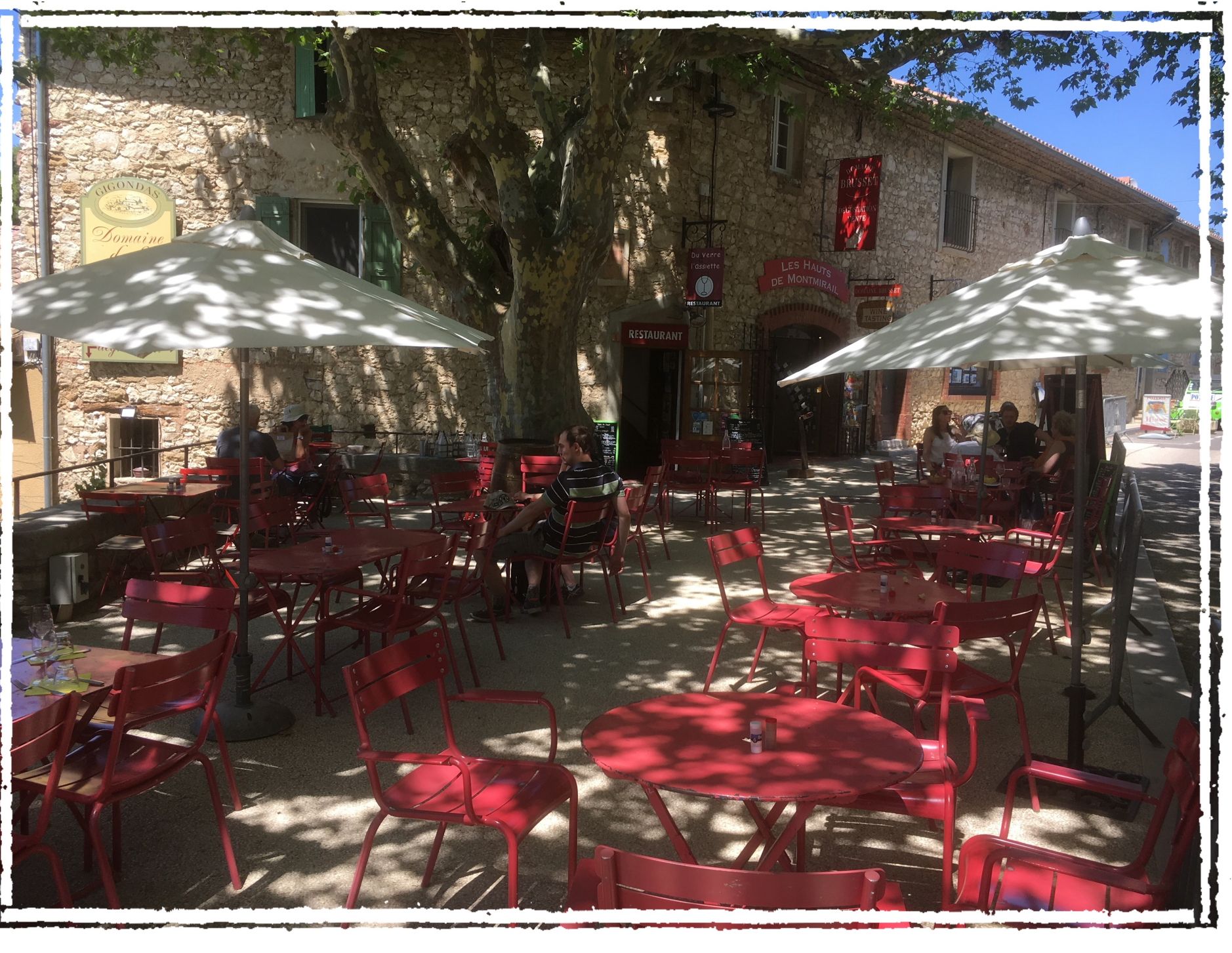 An outdoor cafe with red chairs and tables, stone walls, and umbrellas providing shade.