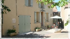 Un edificio con facciata beige, persiane azzurro chiaro, un piccolo balcone e una zona salotto esterna coperta con tavolo e sedie.