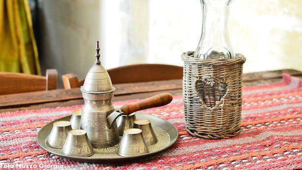 Une théière en métal traditionnelle, dotée d’un bec verseur et d’une anse, un vase en verre et un panier tressé, le tout posé sur une nappe à motifs.