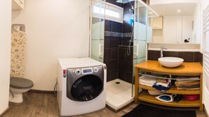 A compact bathroom with a washing machine, a glass shower enclosure, a sink with a bowl, and a wooden shelf with towels. - enjoy this apartment located 10 minutes from the beaches (Cannes)