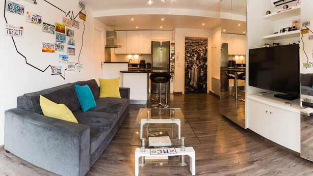 A modern living room with a grey sofa, yellow and blue cushions, a glass coffee table, and a kitchen in the background. - enjoy this apartment located 10 minutes from the beaches (Cannes)