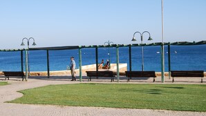 Un parque costero con un camino pavimentado, bancos y una zona cubierta para sentarse con vistas al mar.