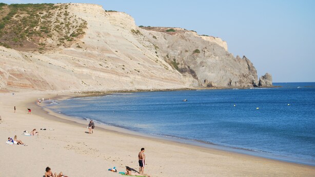 Una playa con gente tomando el sol y nadando, acantilados al fondo y un cielo despejado.