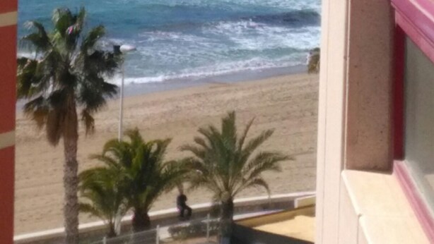 A view of a beach with a mountain in the background, palm trees, and a building with a pink railing.