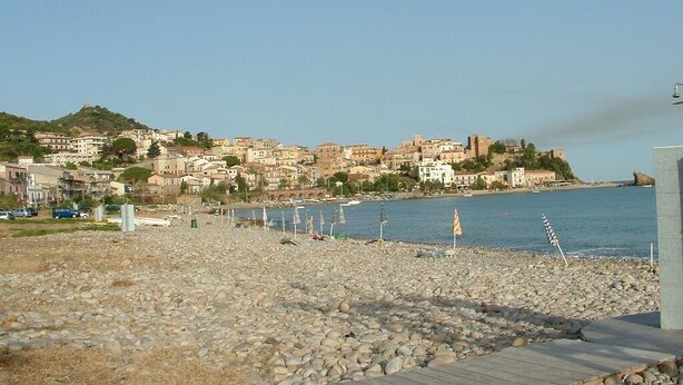 A rocky beach with a wooden walkway, a clear view of a coastal town with buildings and a hill in the background. - Apartment "Full Moon" (Tusa)