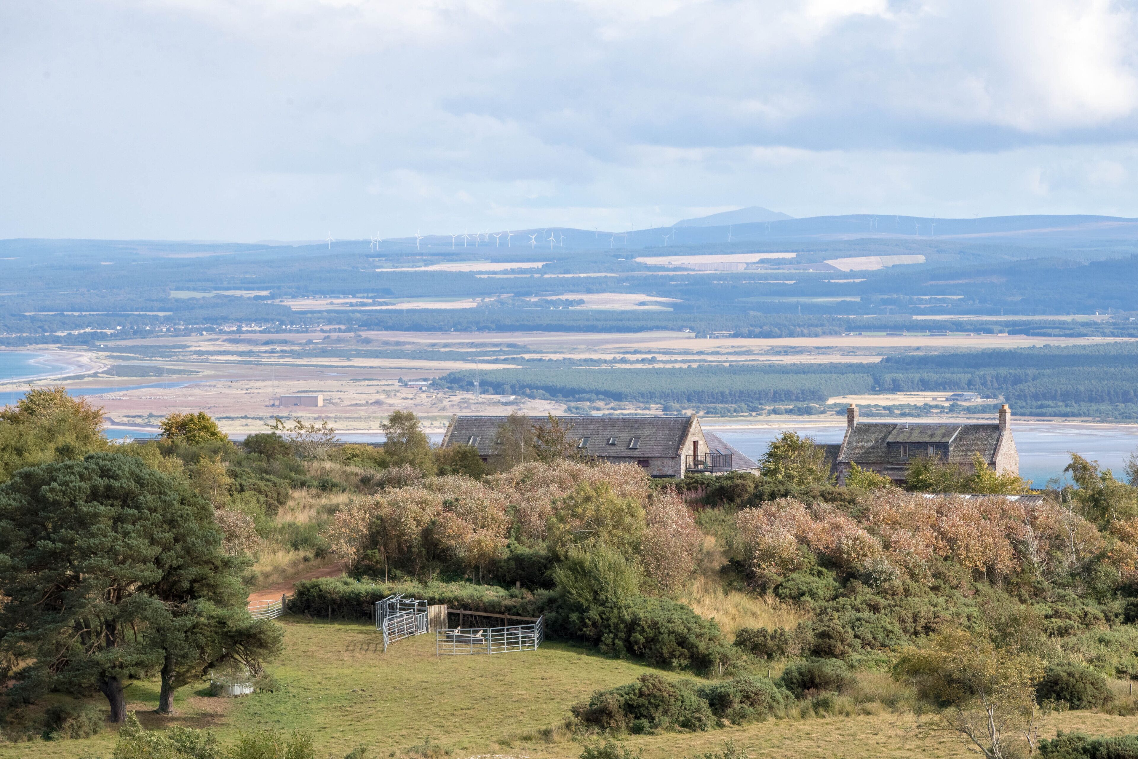 A rural landscape with a stone house, a fenced area, and a distant view of wind turbines and a body of water.