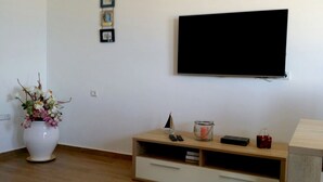 Ein Wohnzimmer mit Flachbildfernseher an der Wand, einem Holz-TV-Schrank mit Fernbedienungen, einer Vase mit Blumen und einem kleinen Schiffmodell.