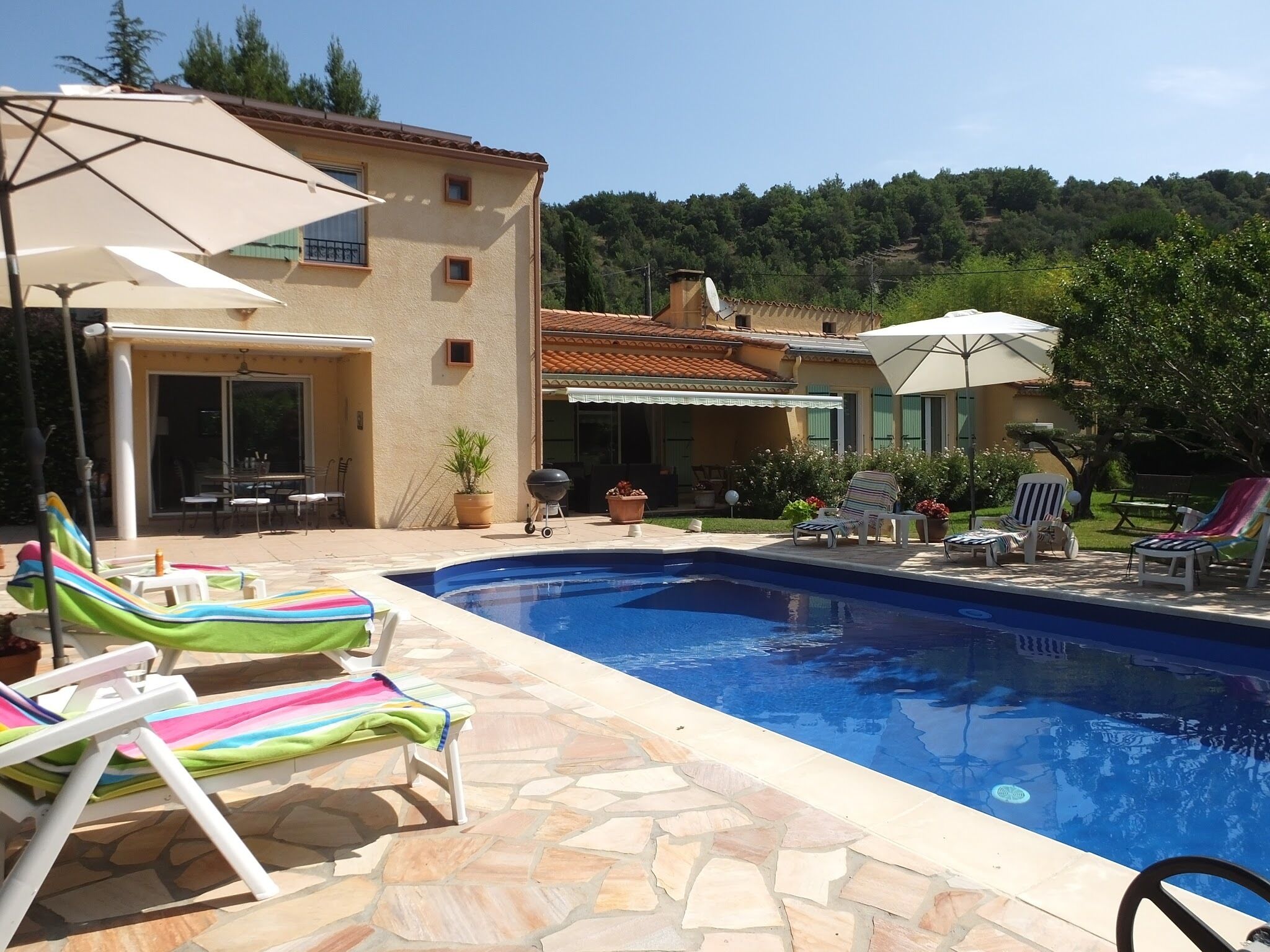 Una piscina con sillones de playa y sombrillas, rodeada por un patio de piedra y una casa con techo de tejas.