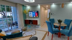 Una sala de estar moderna con televisor, sillones azules, una mesa de centro de vidrio y vistas amplias por un ventanal.