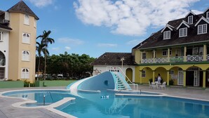 Una piscina con un tobogán azul, rodeada de edificios con balcones y un letrero de restaurante.