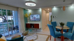 Una sala de estar moderna con televisor, sillas azules, una mesa de centro de vidrio y vista al exterior.