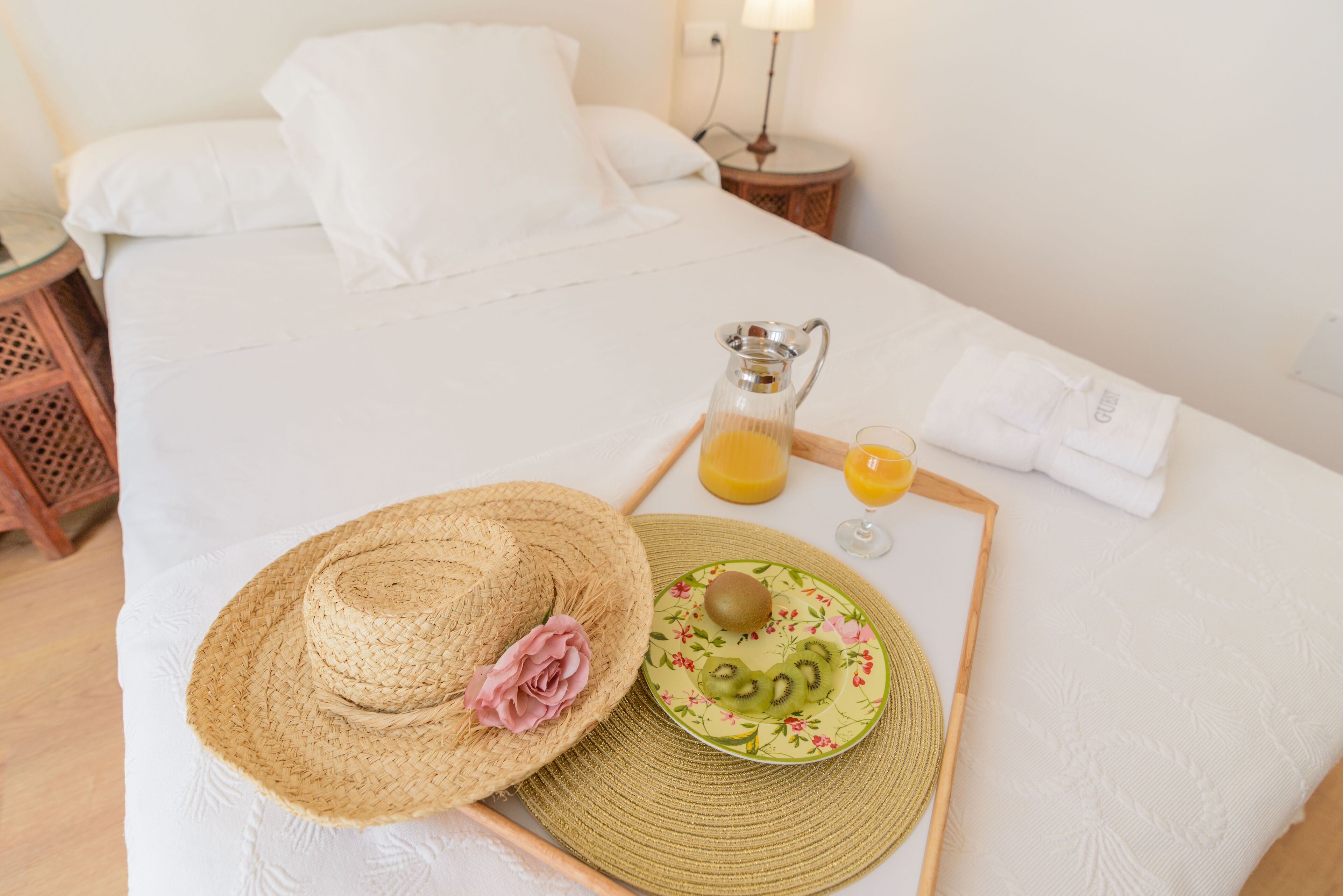 Una cama bien hecha con sábanas blancas, un sombrero de paja con una flor rosa, un plato con rodajas de kiwi, un vaso de jugo de naranja y una jarra de jugo de naranja sobre una bandeja.