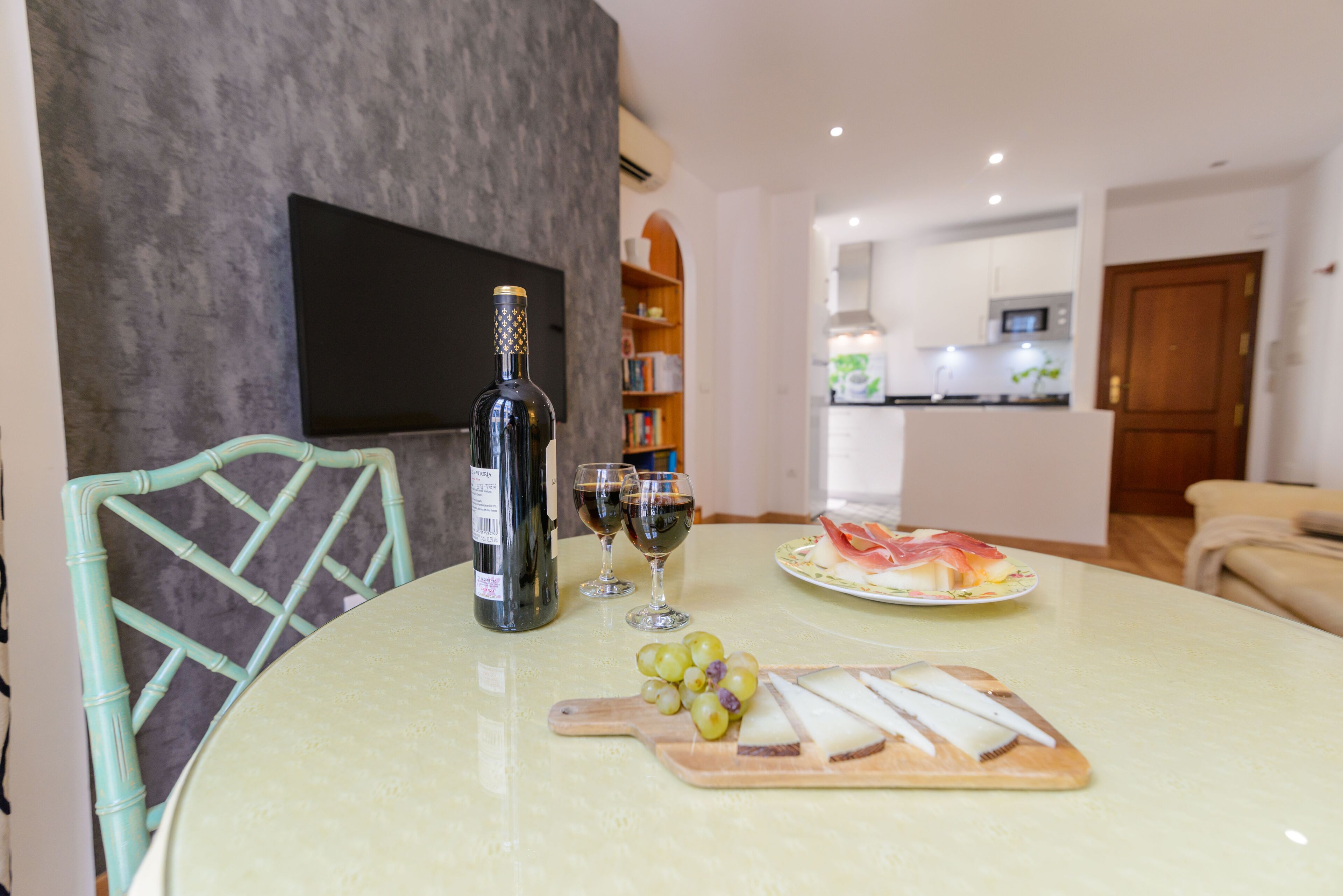 A dining table with a bottle, glasses, grapes, and cheese.