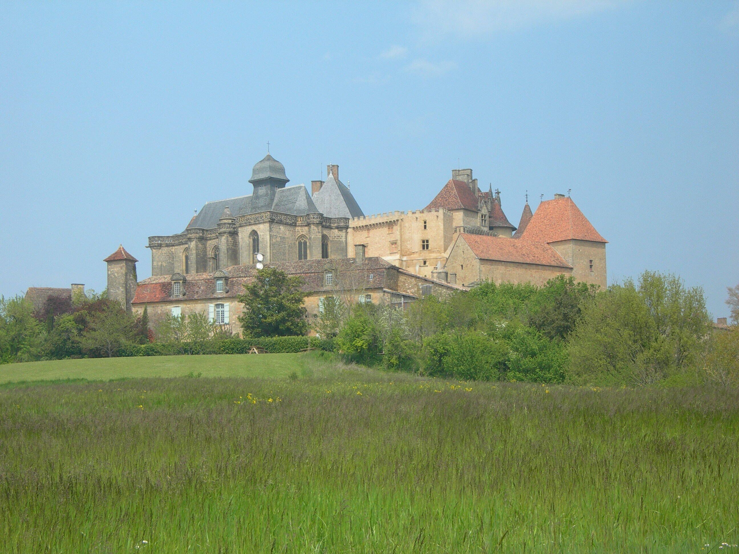 A historic building with a prominent central tower, surrounded by lush greenery and a field.