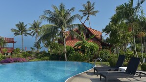 A pool area with lounge chairs, a pool, and a building with a red roof surrounded by palm trees. - 3 bed beach front villa (Koh Samui)