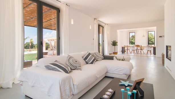 Una sala de estar moderna con un sofá seccional blanco, un comedor con mesa y sillas de madera, y ventanas grandes que ofrecen vistas al exterior.