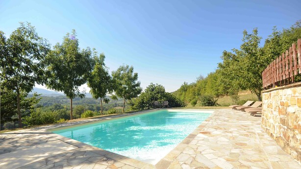 Piscina con baldosas de piedra, rodeada de árboles y un muro de piedra.