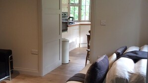 Een woonkamer met een bank, een bijzettafeltje en een stoel. Een keuken is zichtbaar door een open deur.