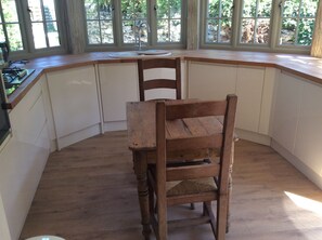 A corner kitchen with a wooden chair, white cabinets, and a window with a view of greenery.