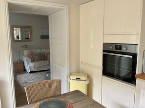 Een functionele keuken met een inbouwoven, een kleine prullenbak en een rieten stoel aan een houten tafel.