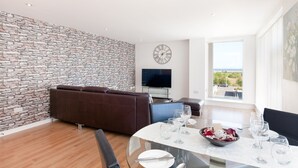 Una sala de estar moderna con una pared de piedra, un sofá de cuero marrón, un televisor de pantalla plana y un comedor con una mesa y sillas de vidrio.