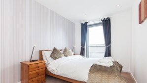 Un dormitorio con cama de madera, ropa de cama blanca, una mesita de noche de madera y una ventana con cortinas.