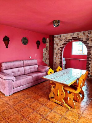 A room with a red wall, a purple sofa, a wooden table with chairs, and a stone wall with a window.