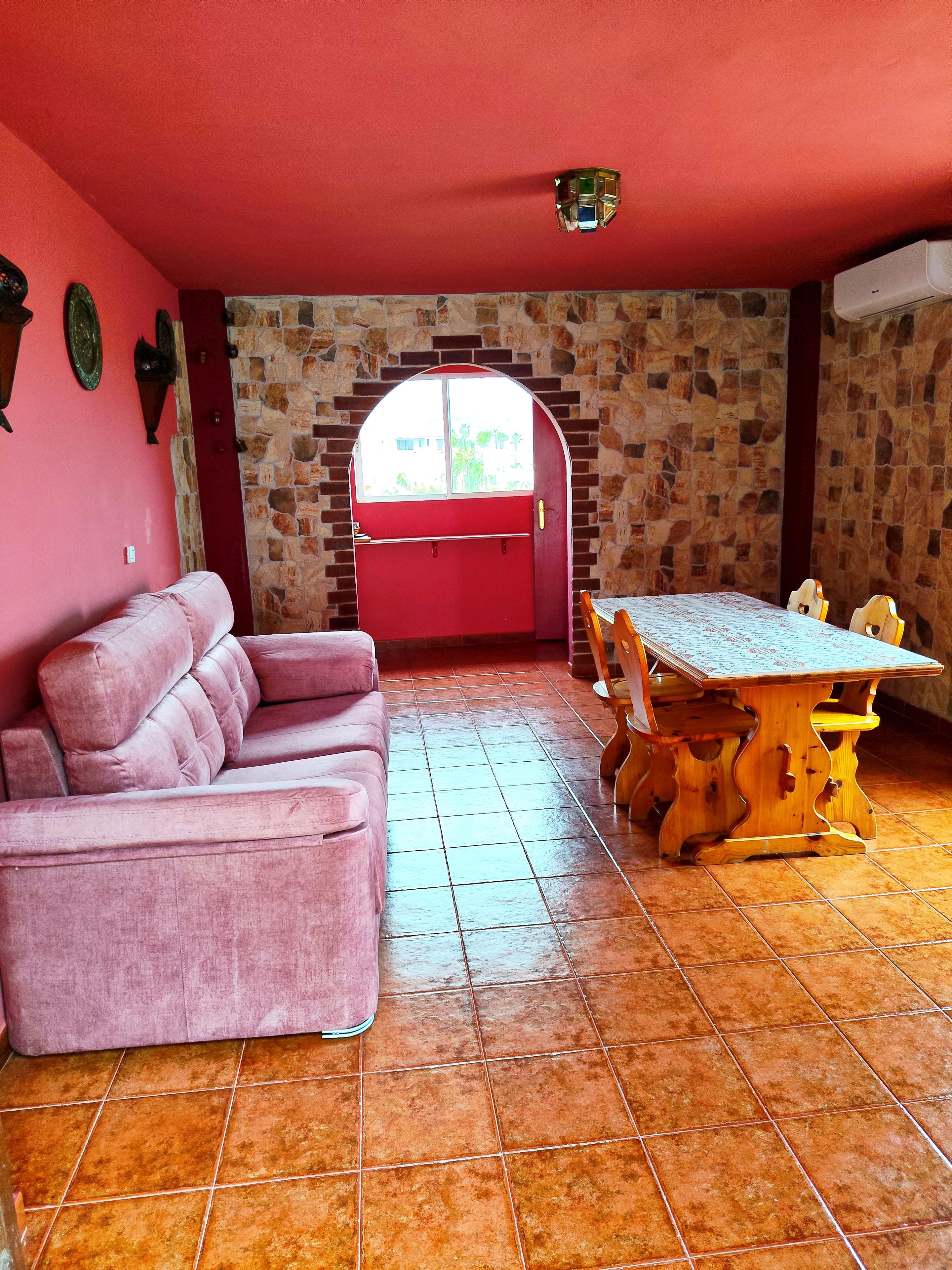 Une pièce avec un mur rouge, un mur en pierre, un canapé rose, une table à manger en bois avec des chaises, et une fenêtre donnant sur un beau paysage.