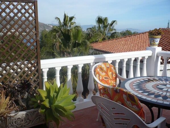 A balcony with a white balustrade, a round table with a mosaic top, and a chair with a patterned cushion.