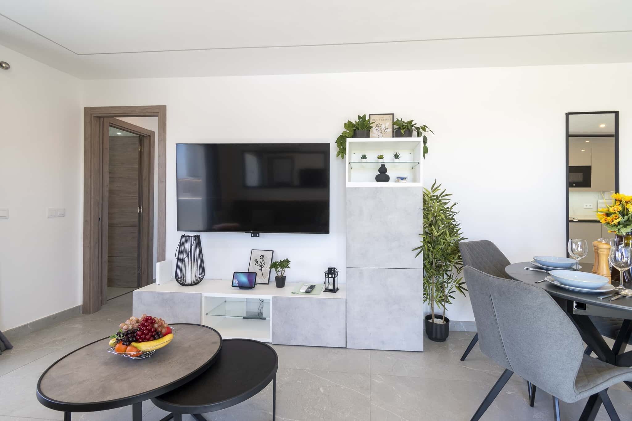 Una sala de estar moderna con un televisor de pantalla plana, un comedor con una mesa lista para dos personas y una zona de estar con una mesa redonda.