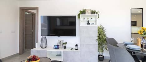 Una sala de estar moderna con un televisor de pantalla plana, un comedor con una mesa lista para dos personas y una zona de estar con una mesa redonda.
